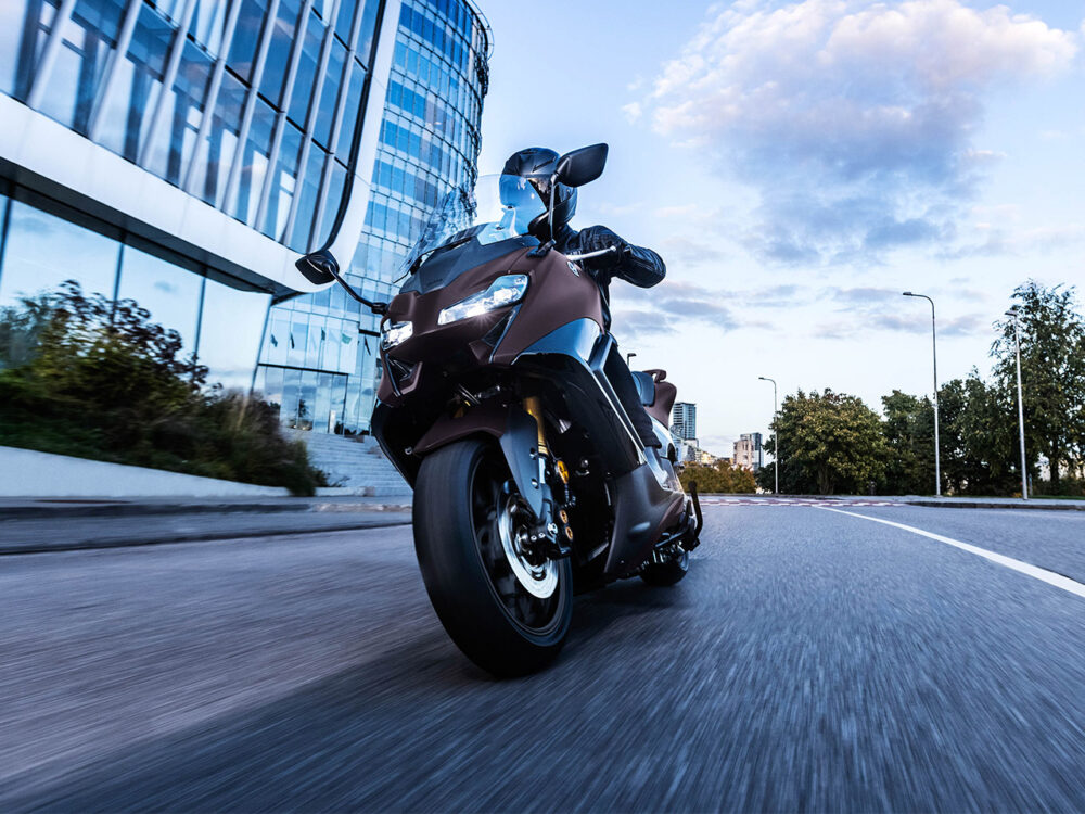 A YAMAHA TMAX 560 - 2024 cruises on an urban road, flanked by sleek glass buildings and trees, beneath a partly cloudy sky.