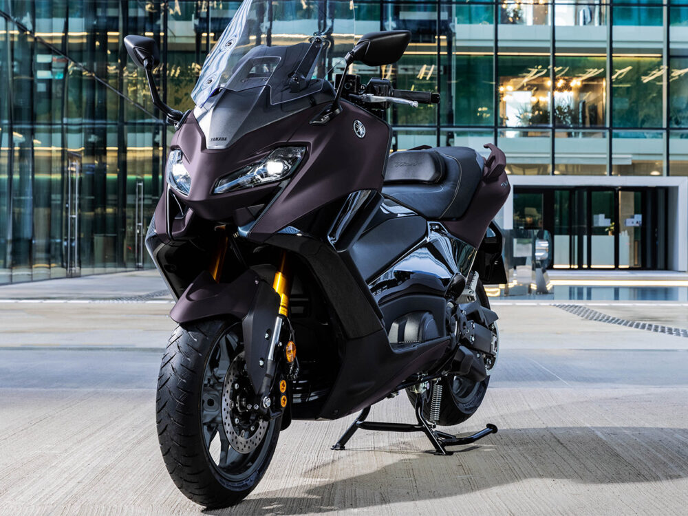 A stylish YAMAHA TMAX 560 - 2024, in glossy black, is parked in front of a modern glass building.