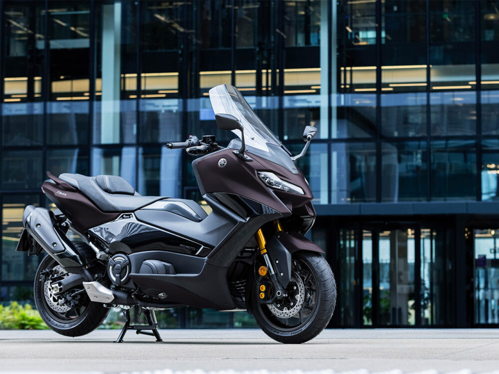 A YAMAHA TMAX 560 - 2024, sporting a sleek dark design, is parked on a pavement in front of a modern glass building; its clear windshield and detailed components are visible.