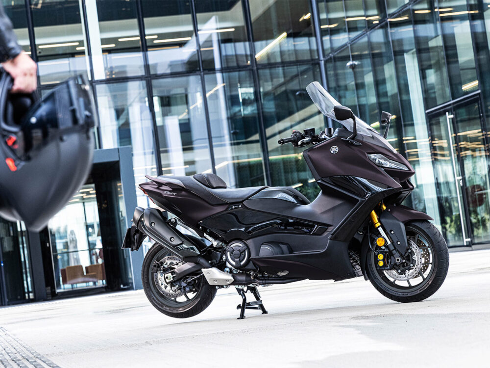 A sleek YAMAHA TMAX 560 - 2024 motorcycle in a dark color is parked outside a modern glass building, with a person holding a helmet visible in the foreground.