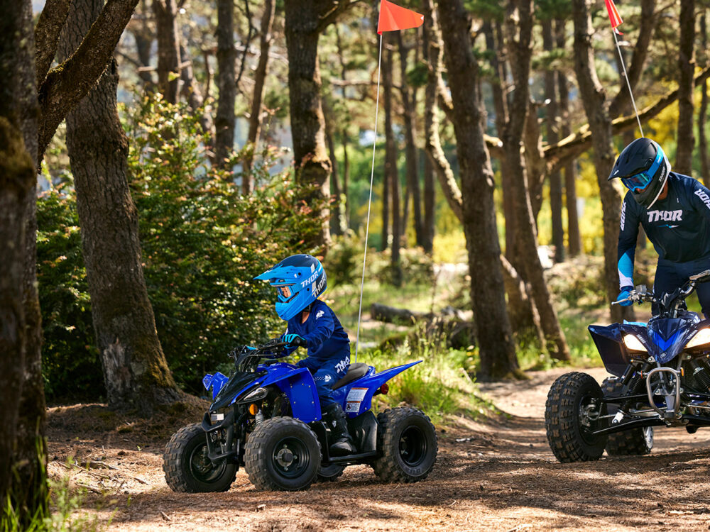 Two people ride YAMAHA YZF50 2025 quad bikes on a forest trail, wearing blue helmets and gear, surrounded by trees and bushes along the dirt path.