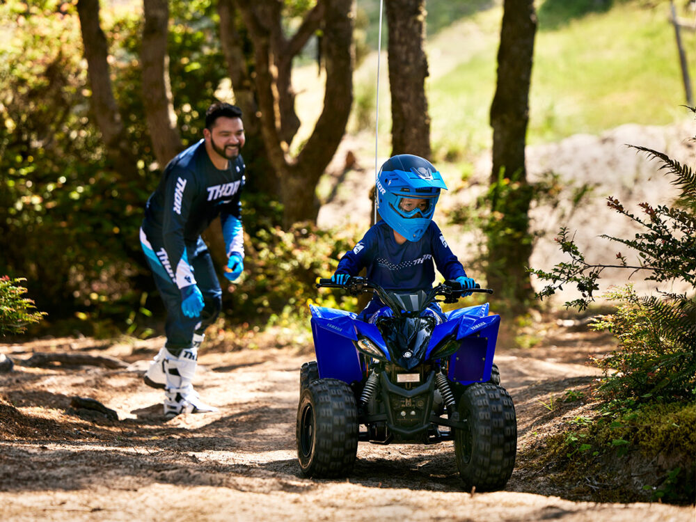 A child rides a YAMAHA YZF50 2025 ATV on a dirt path as an adult in racing gear follows behind in a wooded area.