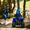 A child rides a YAMAHA YZF50 2025 ATV on a dirt path as an adult in racing gear follows behind in a wooded area.