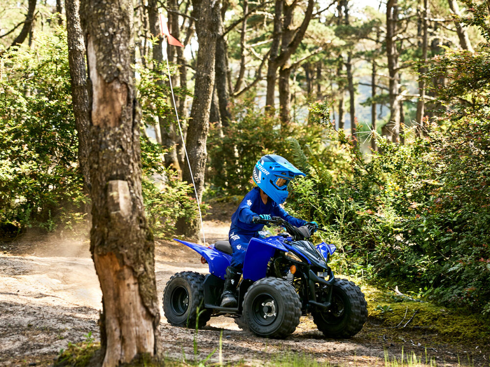 A person is riding a YAMAHA YZF50 2025 ATV through a wooded area, wearing matching blue helmet and protective gear.