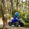 A person is riding a YAMAHA YZF50 2025 ATV through a wooded area, wearing matching blue helmet and protective gear.