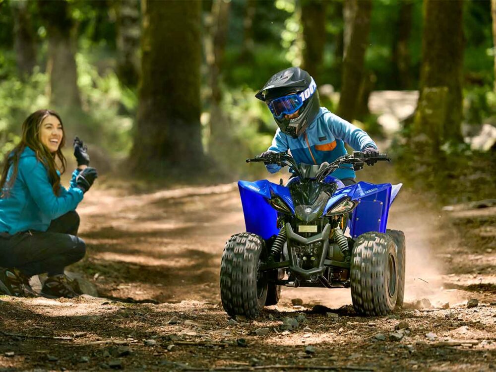 A child navigates through a forest on a YAMAHA YZF50 2025, sporting a helmet and goggles. An adult in blue attire kneels nearby, smiling proudly.