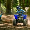 A child navigates through a forest on a YAMAHA YZF50 2025, sporting a helmet and goggles. An adult in blue attire kneels nearby, smiling proudly.