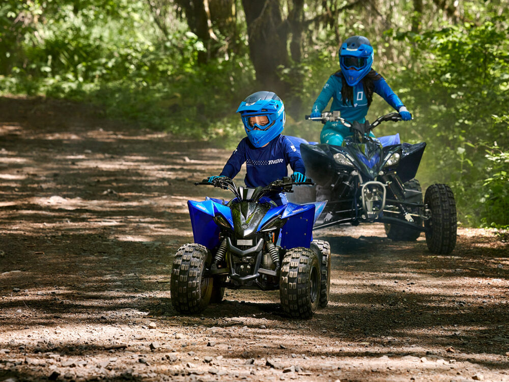 Two individuals on YAMAHA YZF50 2025 ATVs navigate a forest path, enveloped by lush greenery.