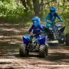 Two individuals on YAMAHA YZF50 2025 ATVs navigate a forest path, enveloped by lush greenery.
