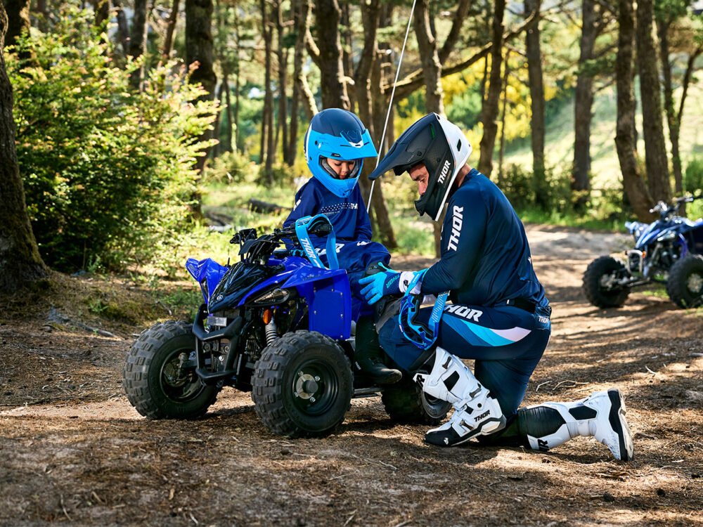 Two people in protective gear are by a small blue YAMAHA YZF50 2025 ATV in a wooded area, with one kneeling and guiding the other.