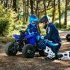Two people in protective gear are by a small blue YAMAHA YZF50 2025 ATV in a wooded area, with one kneeling and guiding the other.