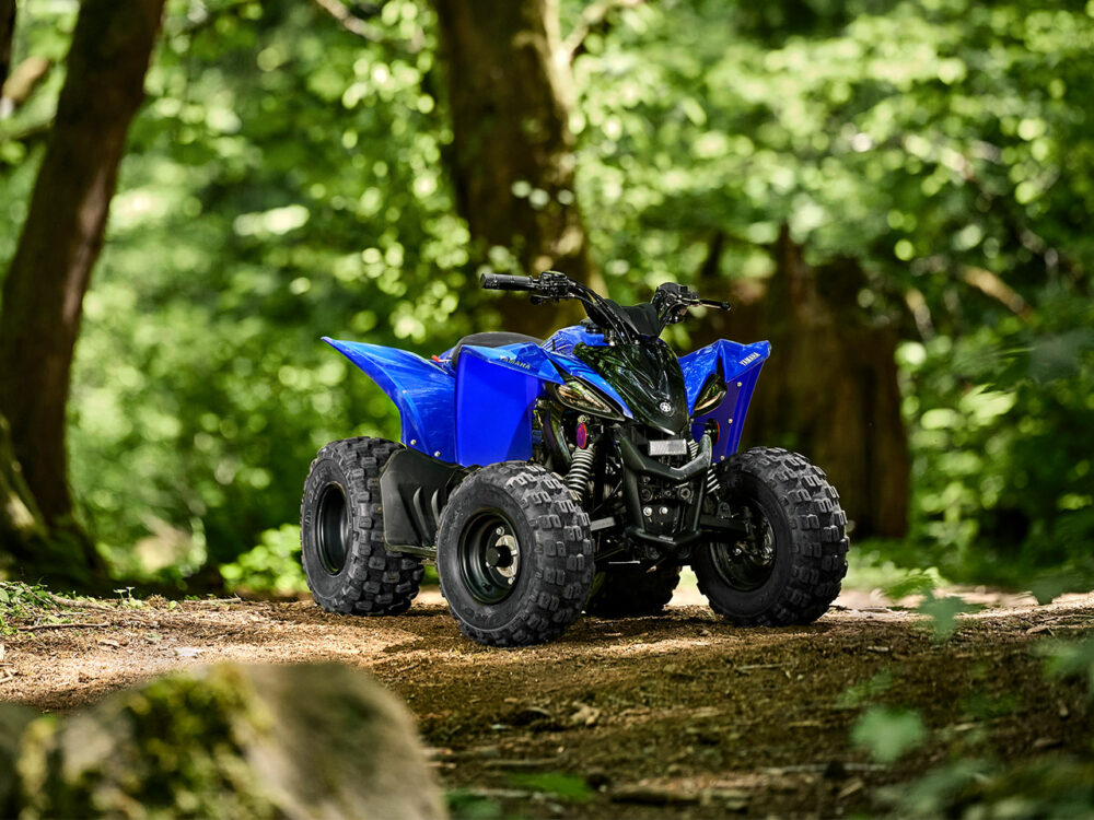 A YAMAHA YZF50 2025 all-terrain vehicle, in blue, is parked on a dirt path amidst a lush green forest.