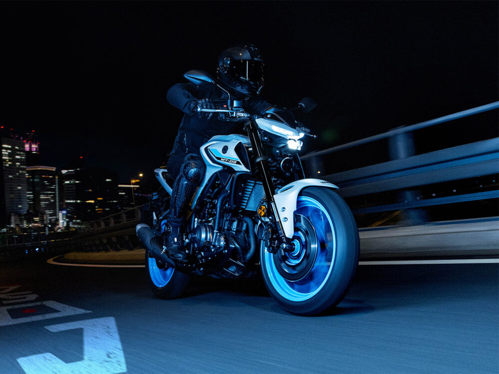 A person rides a YAMAHA MT-03 LAMS 2025 motorcycle with glowing blue wheels on a city road at night, with buildings visible in the background.