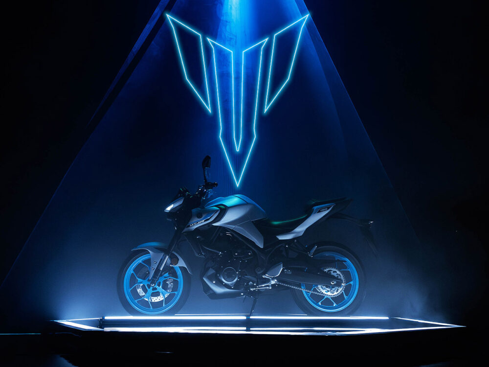 The YAMAHA MT-03 LAMS 2025 is displayed on a platform with glowing blue lights and a triangular neon sign overhead.