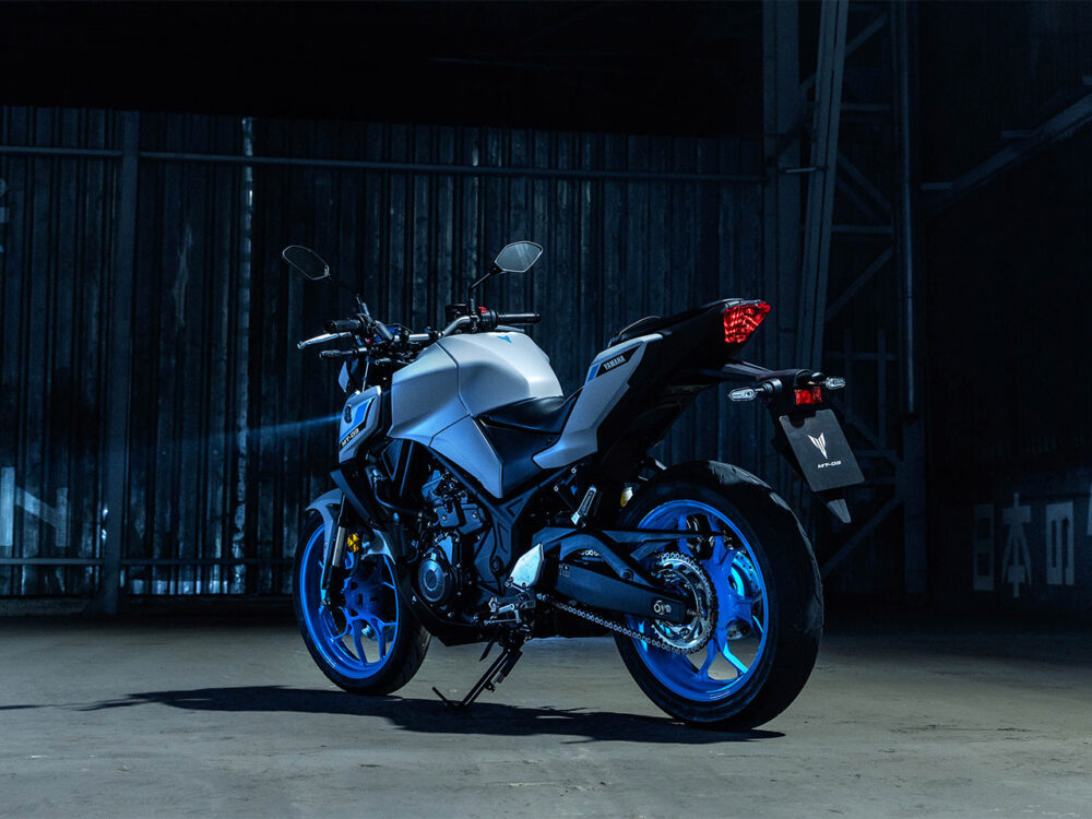 The YAMAHA MT-03 LAMS 2025, featuring a silver body with blue rims, is parked in a dimly lit industrial space, viewed from the rear left side.