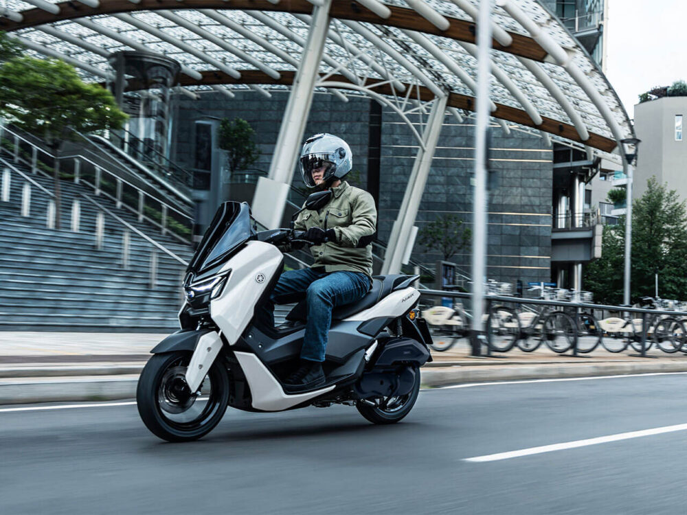 A person is riding a YAMAHA NMAX 155 LAMS scooter on an urban road while wearing a helmet and a green jacket, with a curved architectural structure visible in the background.