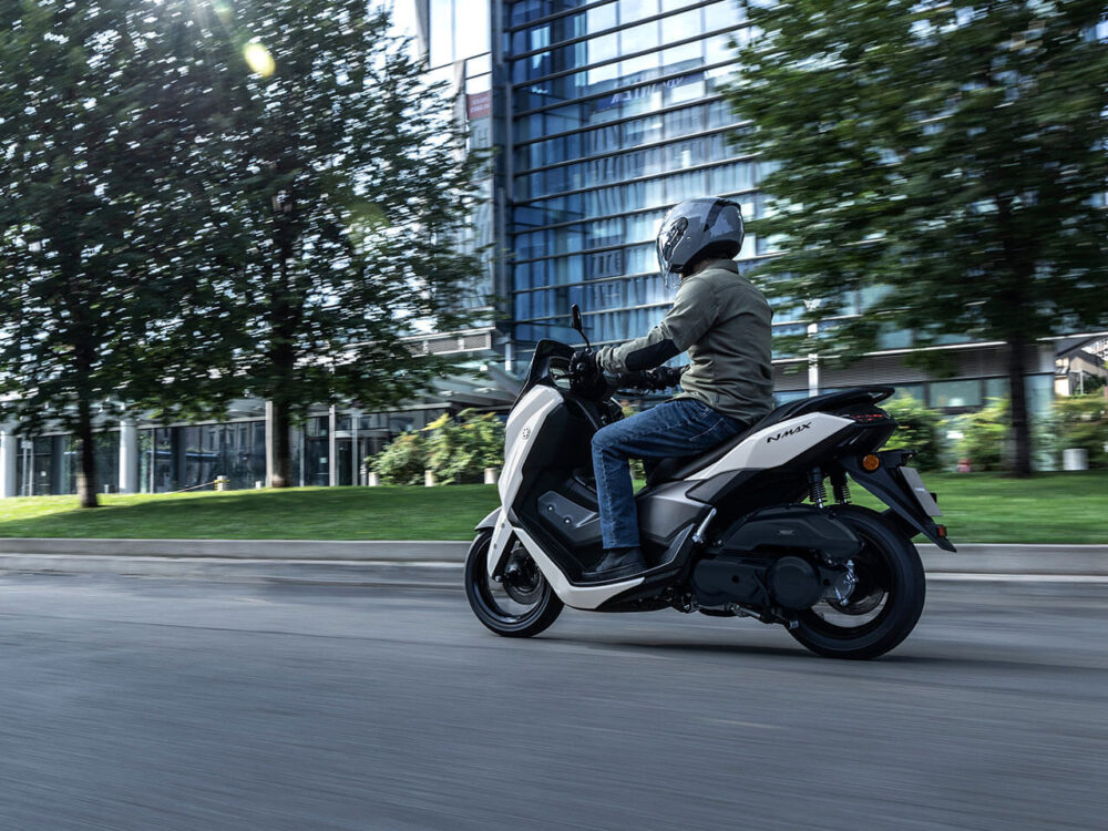 A person wearing a helmet rides a YAMAHA NMAX 155 LAMS scooter on a city street, with buildings and trees in the background.