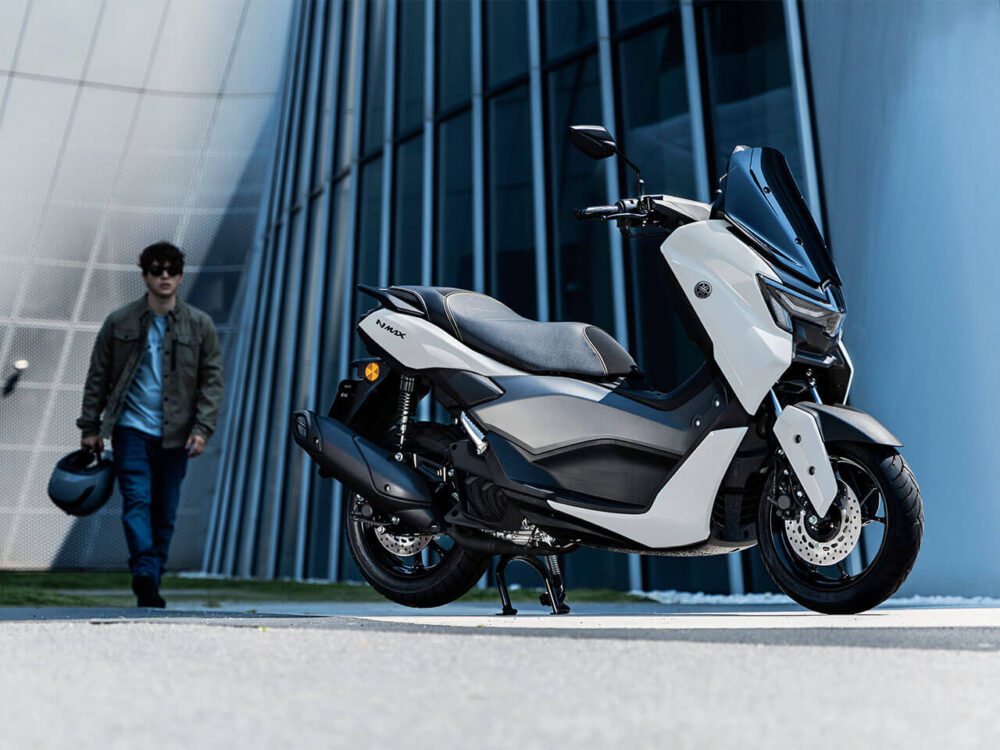 A sleek YAMAHA NMAX 155 LAMS motorcycle in white is parked on a paved area before a modern glass building. A person wearing a jacket walks towards it, holding their helmet.