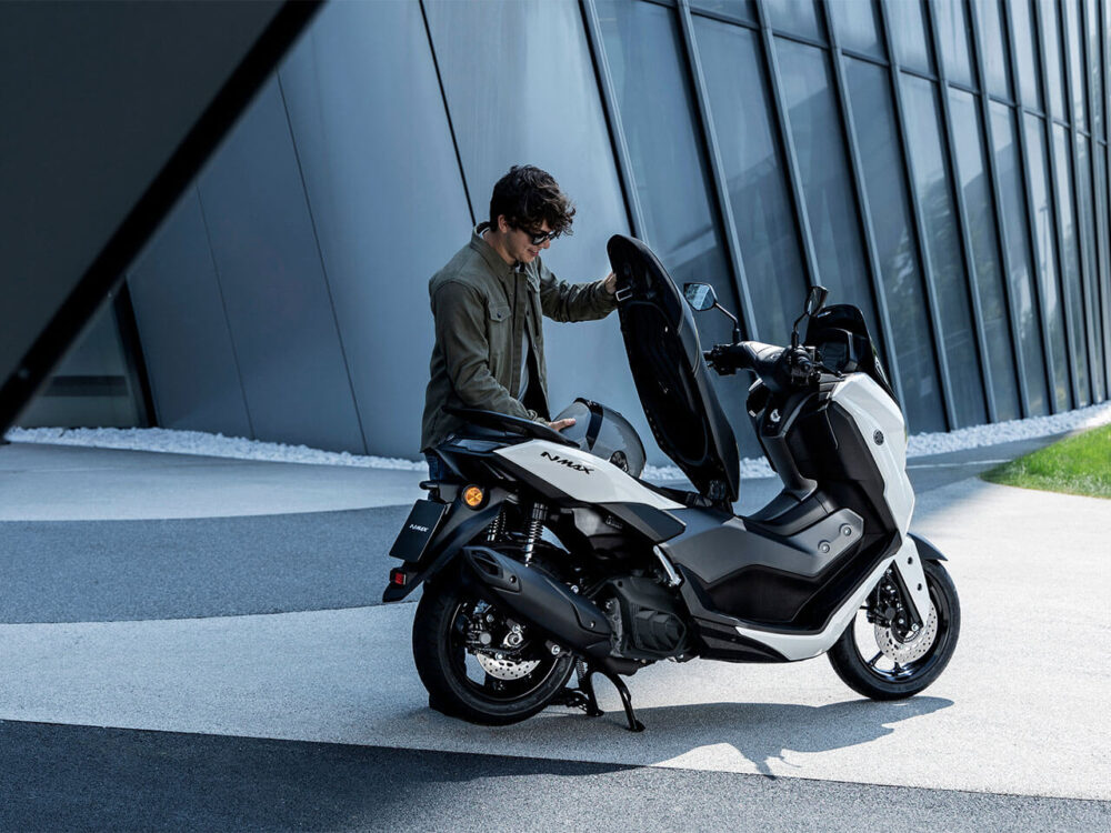 A person in a green jacket examines the under-seat storage of a YAMAHA NMAX 155 LAMS, a white and black scooter, parked on a paved surface near a modern building.