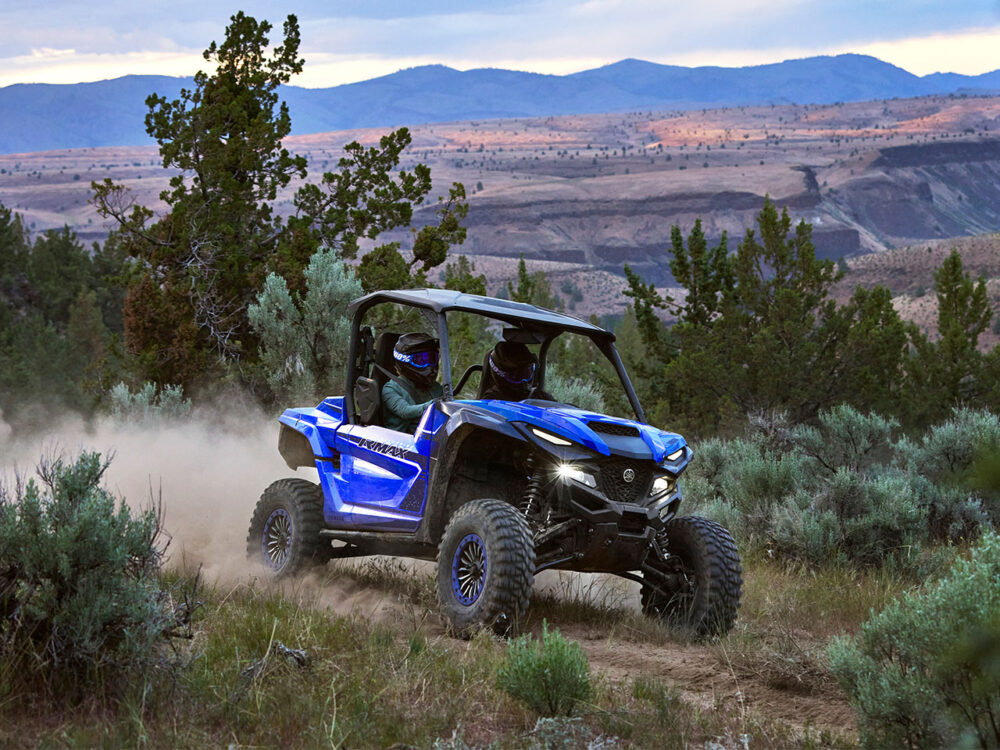 Two people ride a blue YAMAHA WOLVERINE RMAX2 1000 SPORT on a dirt path through hilly terrain with shrubs and mountains under a cloudy sky.