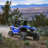 Two people ride a blue YAMAHA WOLVERINE RMAX2 1000 SPORT on a dirt path through hilly terrain with shrubs and mountains under a cloudy sky.