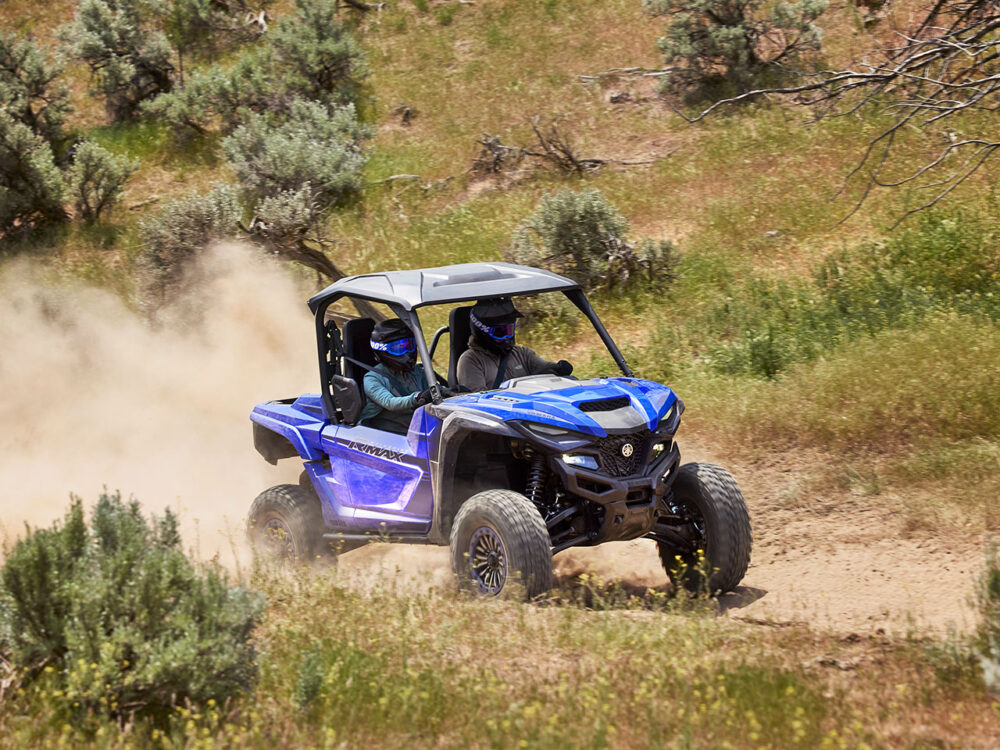 Two helmeted riders navigate a dusty trail surrounded by grass and shrubs in a Yamaha Wolverine RMAX2 1000 Sport, an off-road vehicle.