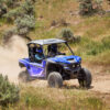 Two helmeted riders navigate a dusty trail surrounded by grass and shrubs in a Yamaha Wolverine RMAX2 1000 Sport, an off-road vehicle.