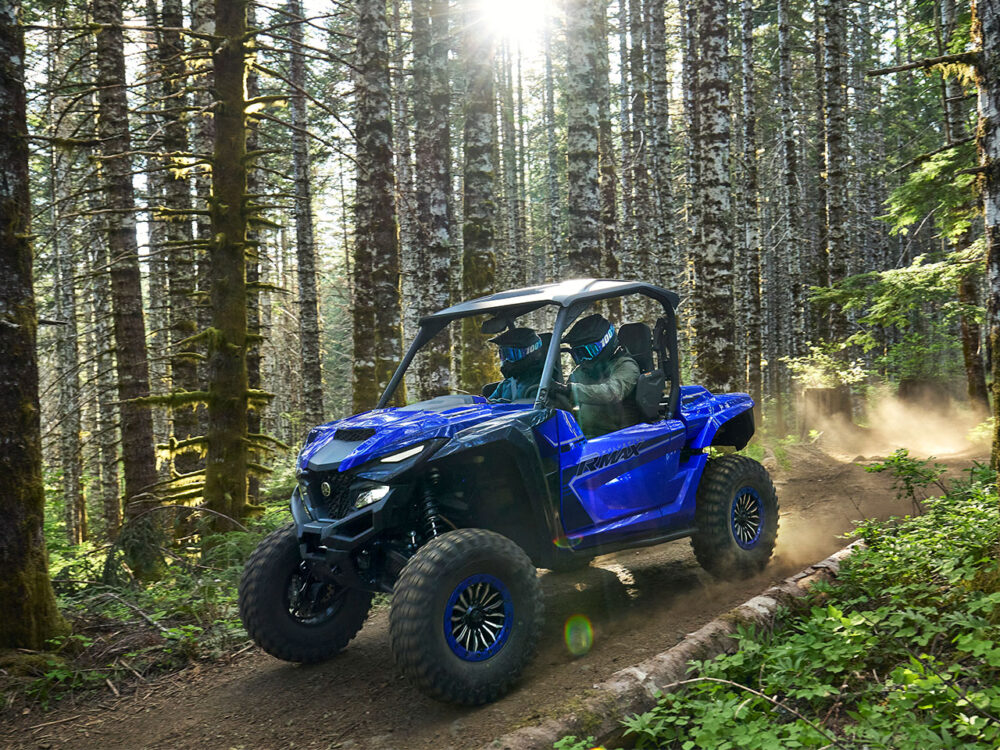 A YAMAHA WOLVERINE RMAX2 1000 SPORT navigates a dense forest trail, kicking up dust as sunlight filters through the trees.