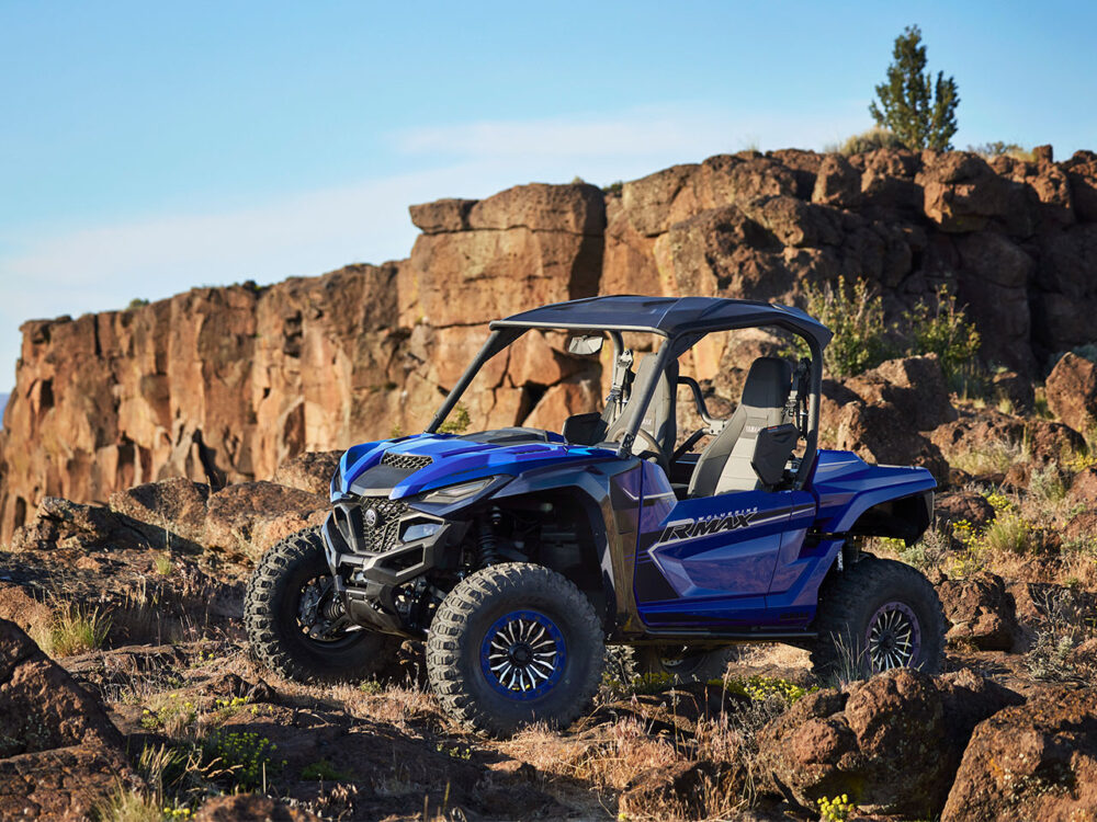 A YAMAHA WOLVERINE RMAX2 1000 SPORT in blue is parked on rocky terrain with rugged cliffs and a clear sky in the background.