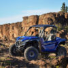 A YAMAHA WOLVERINE RMAX2 1000 SPORT in blue is parked on rocky terrain with rugged cliffs and a clear sky in the background.