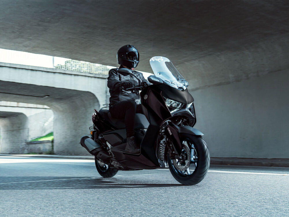 A person rides a black YAMAHA XMAX 300 LAMS 2025 scooter under a highway overpass, wearing a helmet and dark clothing in daylight.