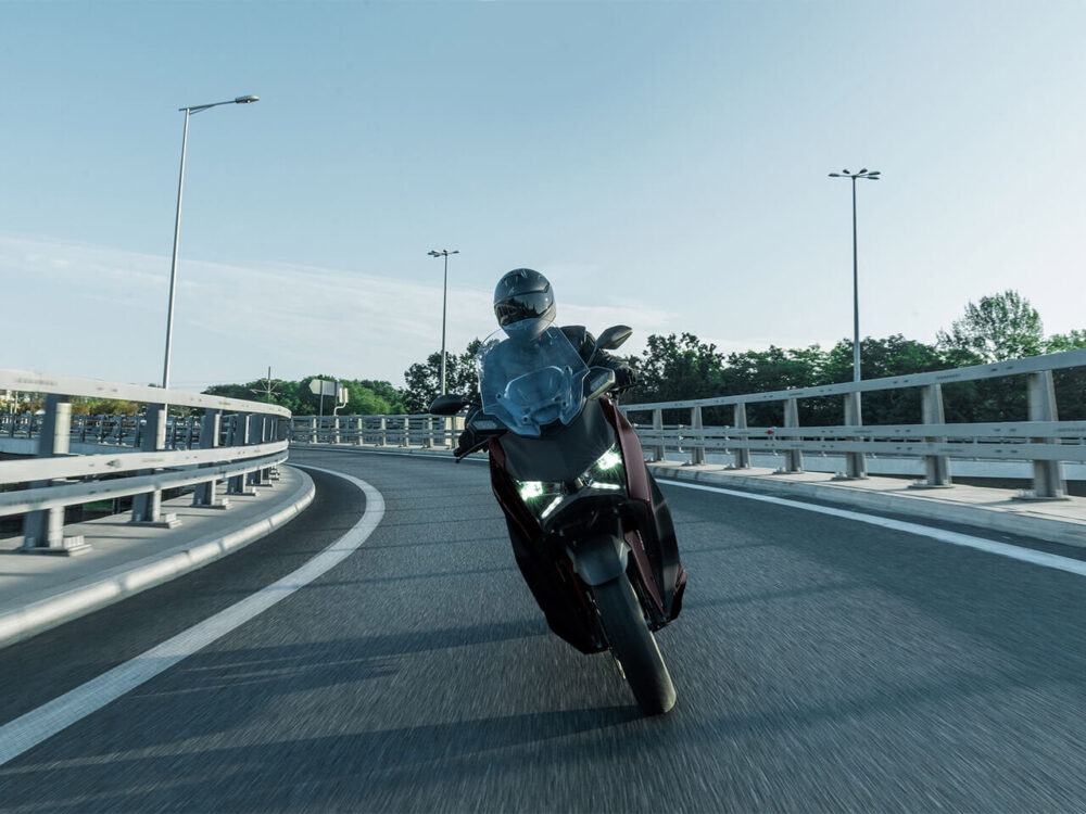 A motorcyclist rides the YAMAHA XMAX 300 LAMS 2025 through a highway curve, with trees and streetlights in the background.