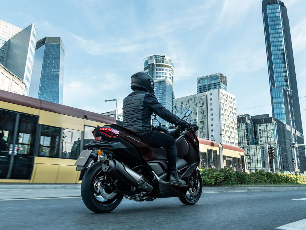 Someone cruises down a city street on the 2025 YAMAHA XMAX 300 LAMS, flanked by soaring skyscrapers and a passing tram.