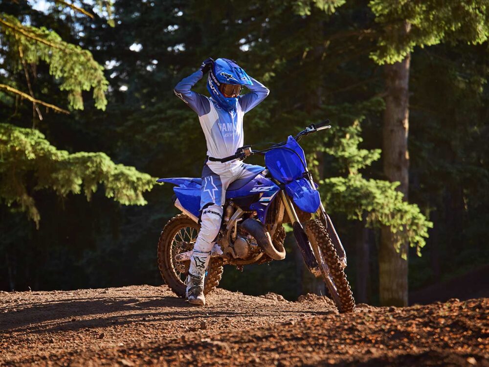 A rider on a YAMAHA YZ125 2025 adjusts their helmet in a sun-dappled forest.