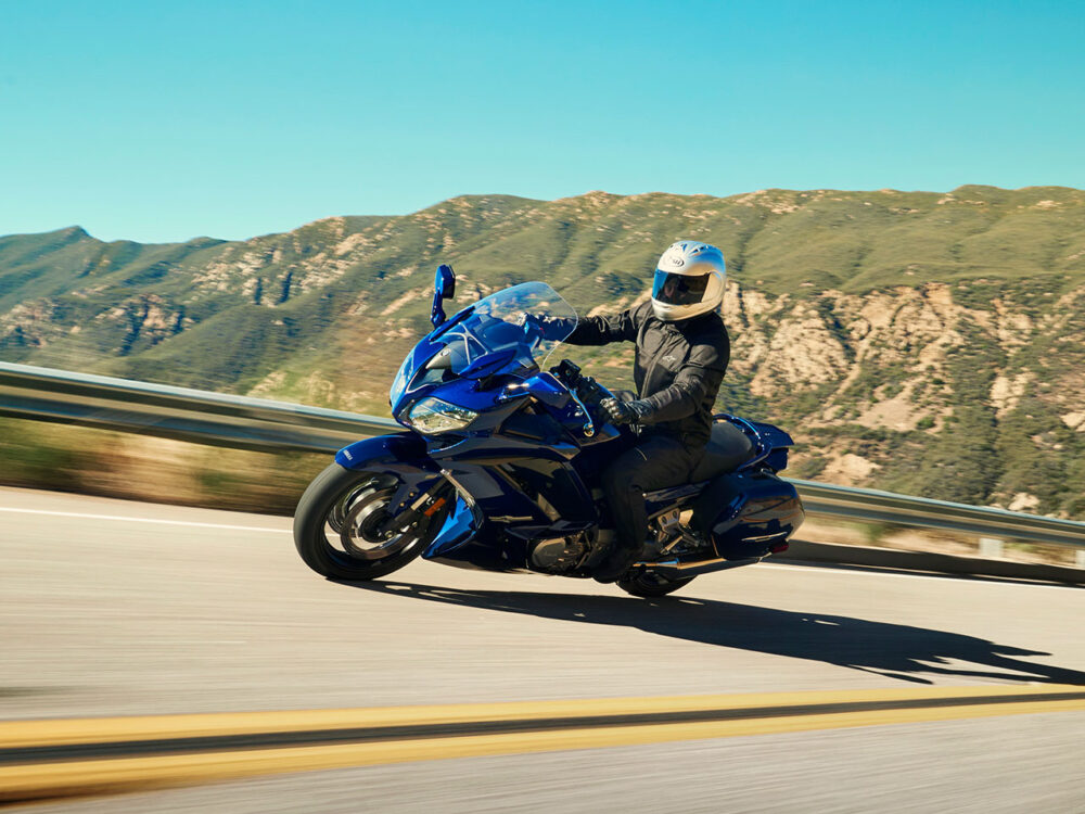 A rider in protective gear navigates a blue YAMAHA FJR 1300 2024 on a winding mountain road, set against a backdrop of clear sky and hills.