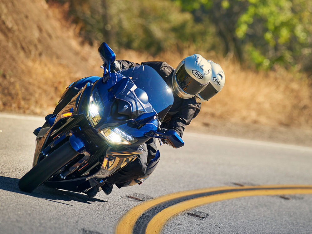 Two individuals in helmets lean into a curve on the road with yellow lines while riding the blue YAMAHA FJR 1300 2024 motorcycle.