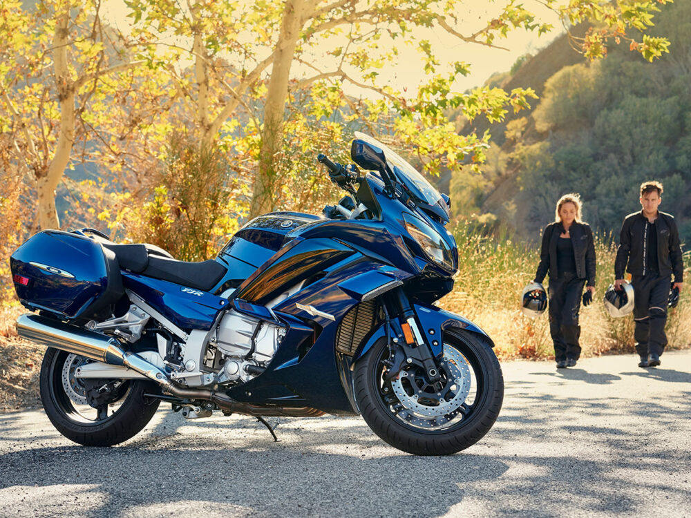 A YAMAHA FJR 1300 2024 is parked on a sunlit road. In the background, two people walk towards it, each holding a helmet. Trees with yellow leaves are on either side.