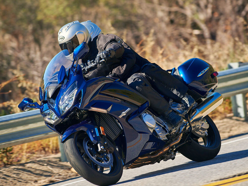 A person wearing a helmet rides a YAMAHA FJR 1300 2024, a blue motorcycle, around a curve on a road with a guardrail, against a blurred background.