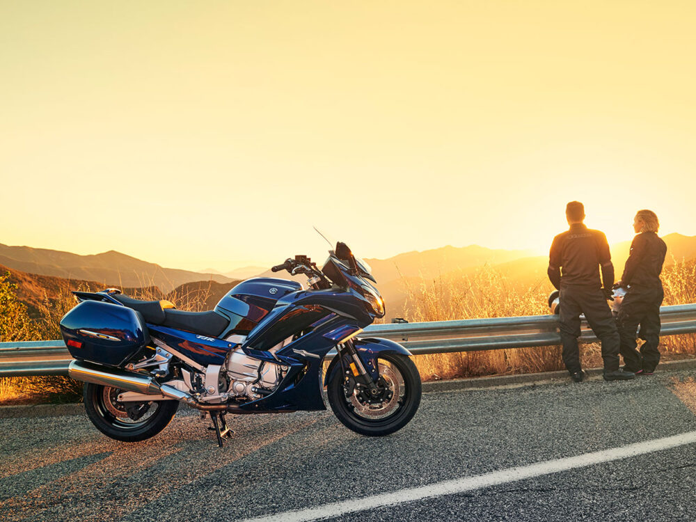 Two people stand by a guardrail, watching the sunset, with a YAMAHA FJR 1300 2024 blue motorcycle parked nearby on the roadside.