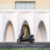 The CFMOTO 150SC is parked in front of a yellow building with white arched accents.