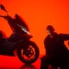 A person sits beside a CFMOTO 150SC scooter against a vivid red backdrop, holding a helmet.