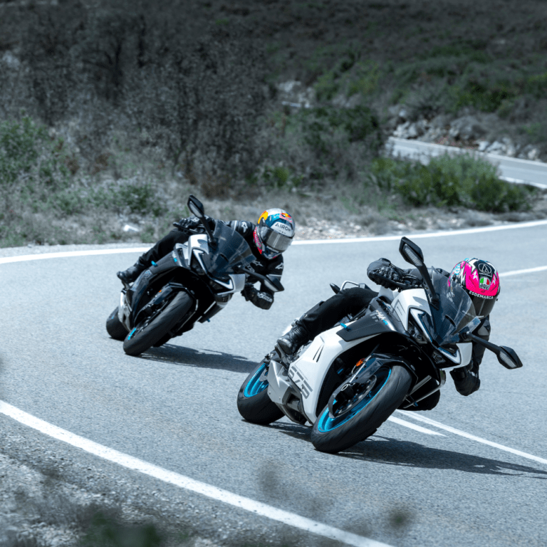 Two motorcyclists lean into a sharp turn on a winding road, showcasing dynamic riding skills and high-speed action.