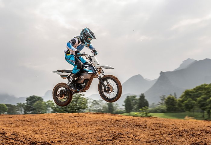 A motocross rider in full gear launches a CX-2E dirt bike off a ramp on a rugged track, with mountains and trees painting the backdrop.