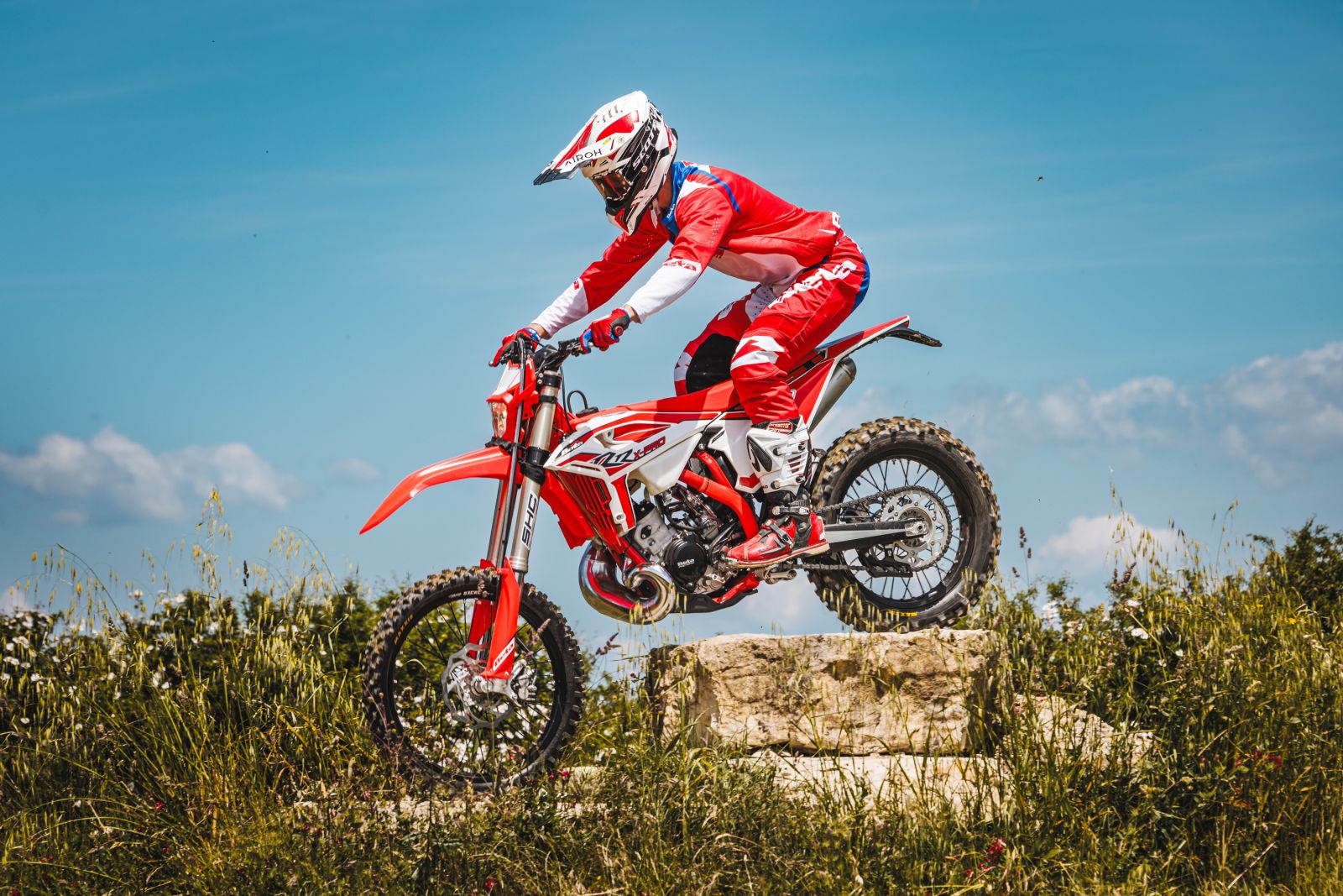 Rider in red gear performs a jump on a red Beta motorcycle over a grassy, rocky terrain under a clear blue sky, unleashing adventure at its finest. Find your thrill with Beta Motorcycles at Trooper Lu Motorcycles.