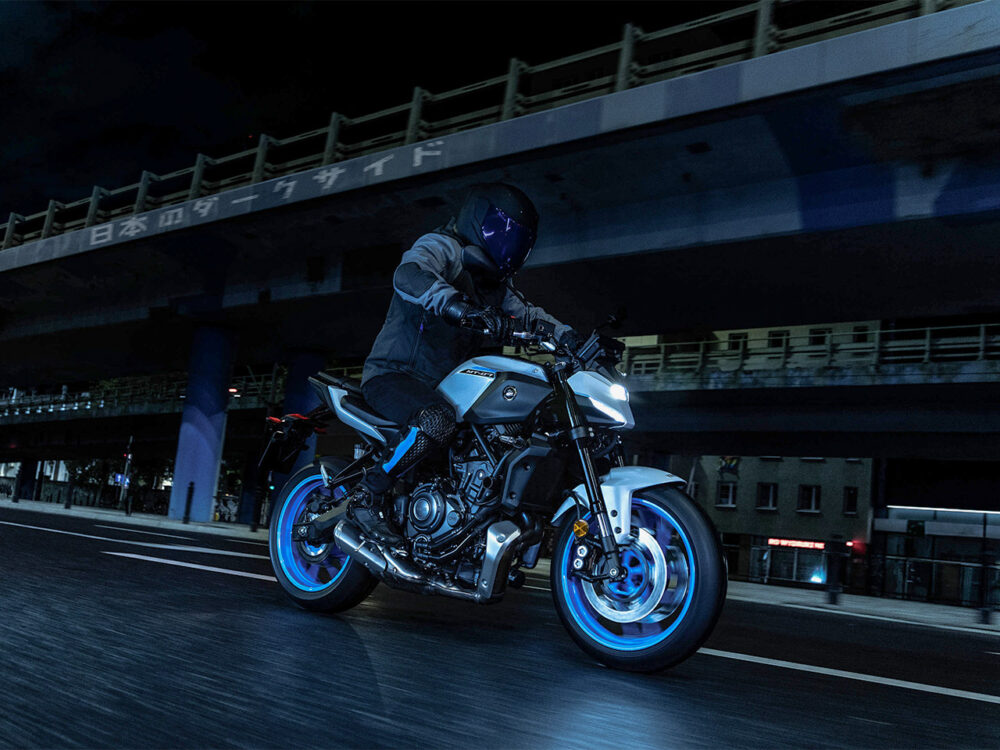 A person rides a YAMAHA MT-07 HO Y-AMT with glowing blue wheels on an urban street at night under an elevated highway.
