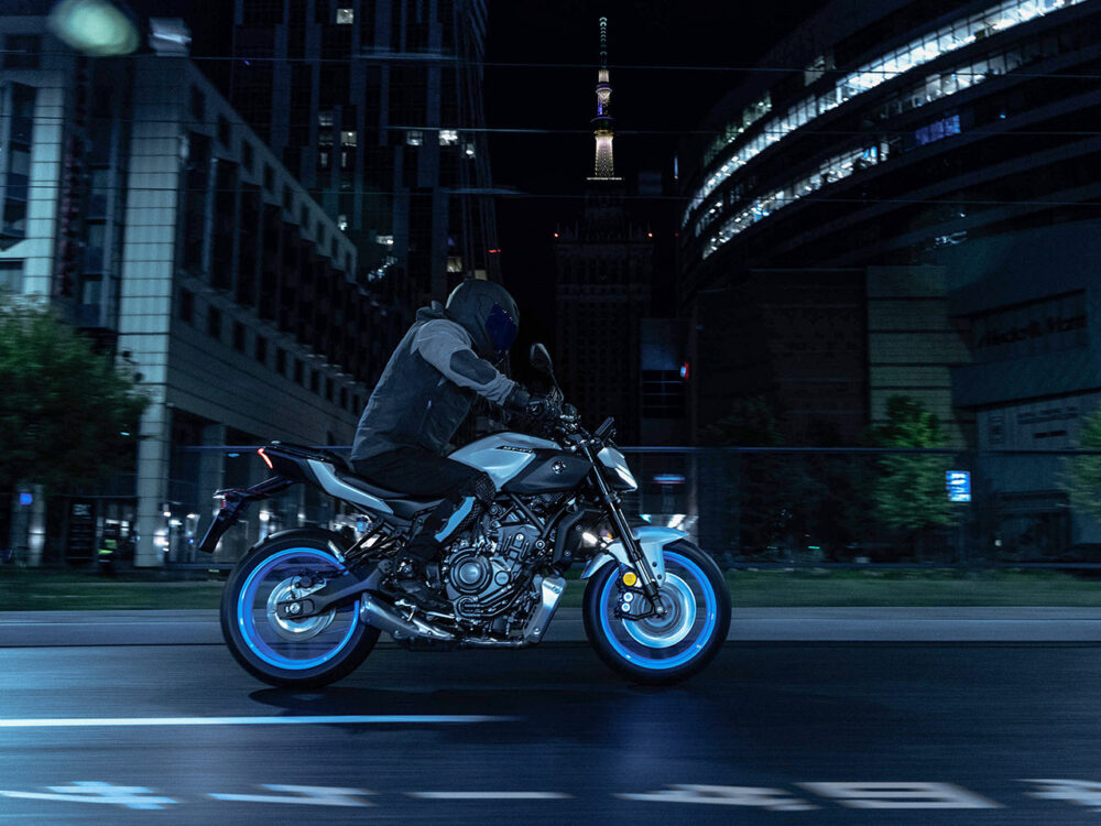 A motorcyclist rides a YAMAHA MT-07 HO Y-AMT, a blue and gray sports bike, through a city street at night with buildings and a tall illuminated tower in the background.