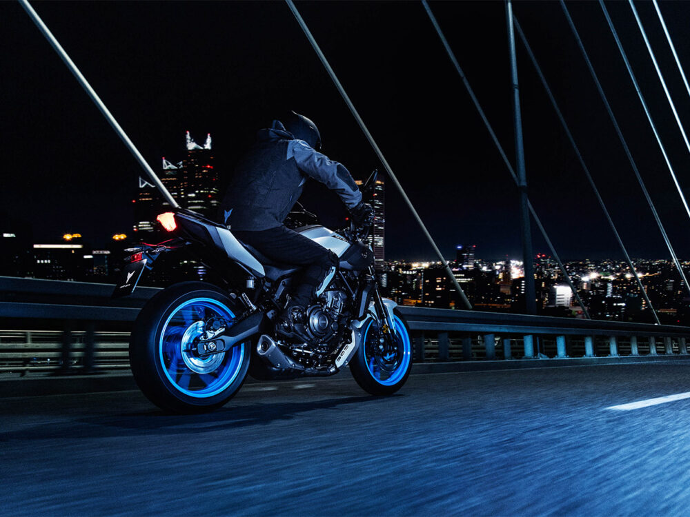 A motorcyclist wearing a helmet rides a YAMAHA MT-07 HO Y-AMT motorcycle with blue wheels across a bridge at night, with a city skyline in the background.