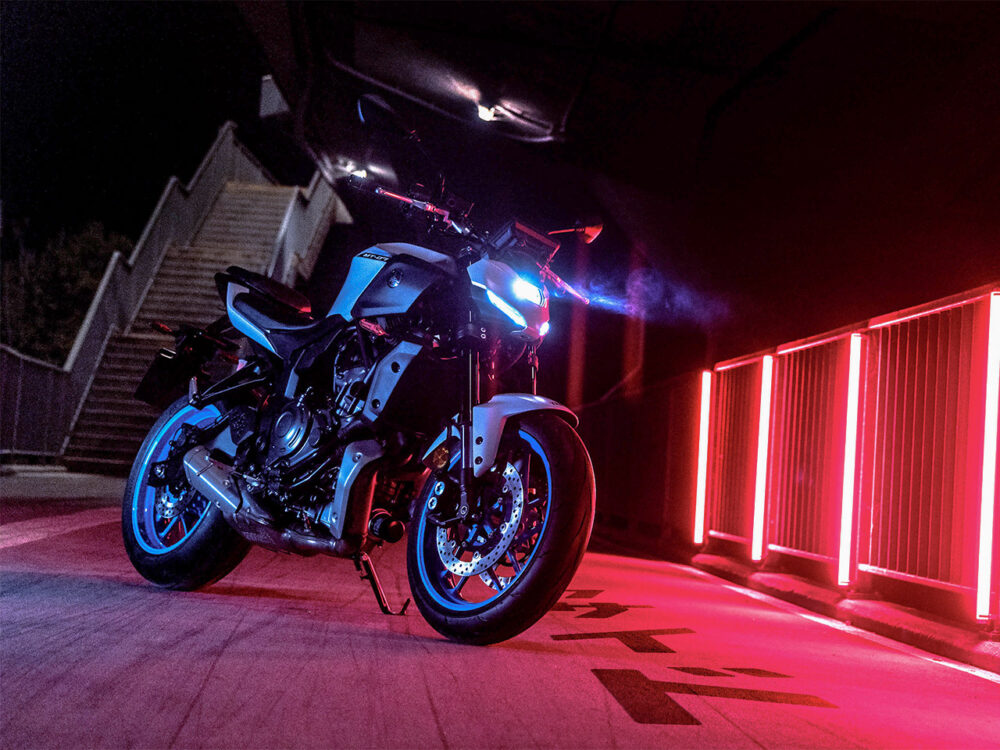 A YAMAHA MT-07 HO Y-AMT motorcycle sits under a bridge at night, lit by red neon lights. Its headlights beam, with stairs visible in the background.