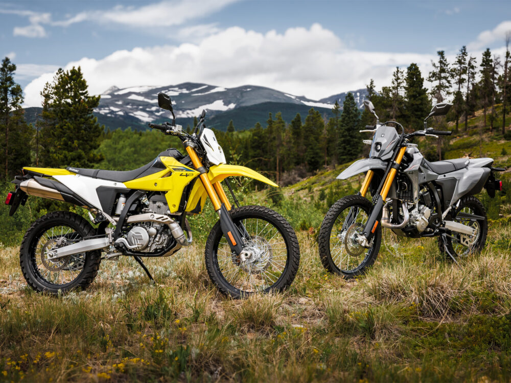 Two SUZUKI DR-Z 4S 2025 dual-sport motorcycles, one yellow and one gray, parked on grassy terrain with mountains and trees in the background.