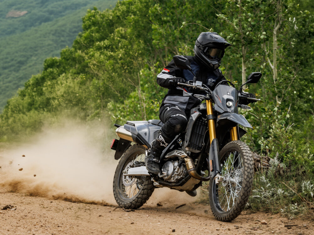 Motorcyclist riding off-road on a trail bike, kicking up dust amidst lush green surroundings.